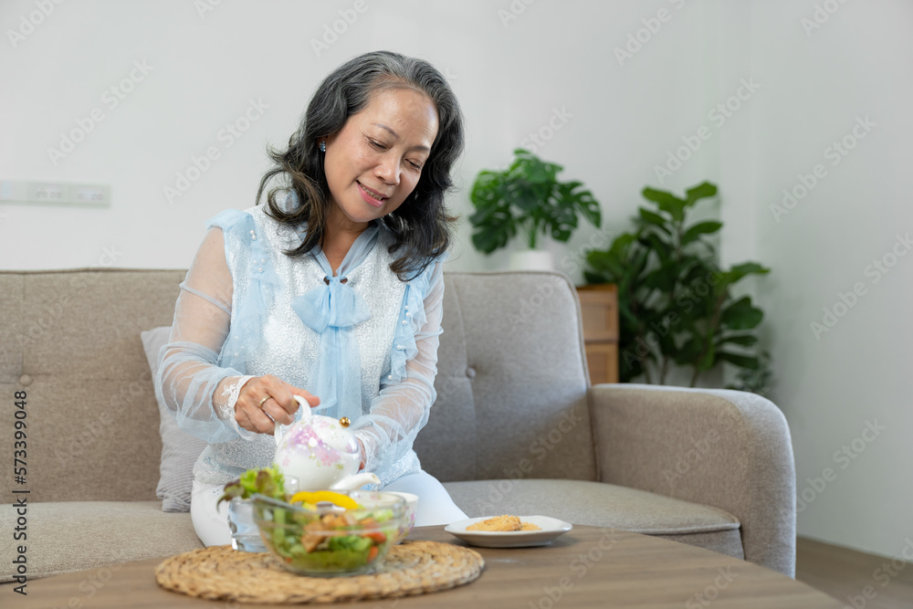 Happy Asian retired woman enjoying her morning tea in the house An elderly woman relaxingly pours and sips hot tea or coffee on the sofa inside her house. Happy Asian retired woman enjoying her morning tea in the house An elderly woman relaxingly pours and sips hot tea or coffee on the sofa inside her house.