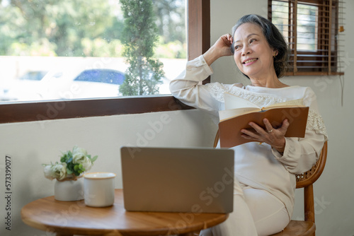 Happy Asian retired woman enjoying reading Books and notes on the subject of health care with a laptop on the table in her morning home in a relaxed and happy way.
