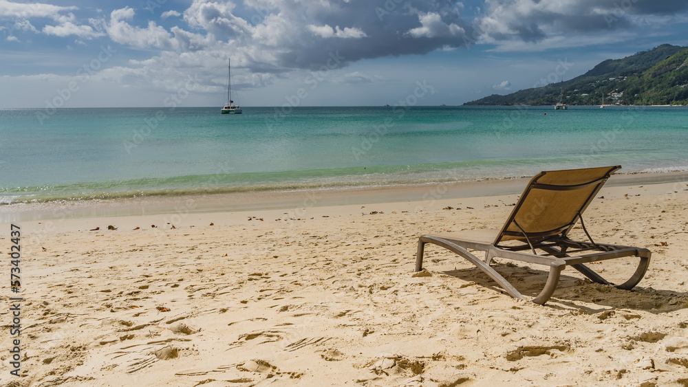 A chaise longue stands on a tropical beach. Footprints in the sand. The waves of the turquoise ocean roll ashore. Yachts in the distance. A green hill against a blue sky and clouds. Seychelles. 