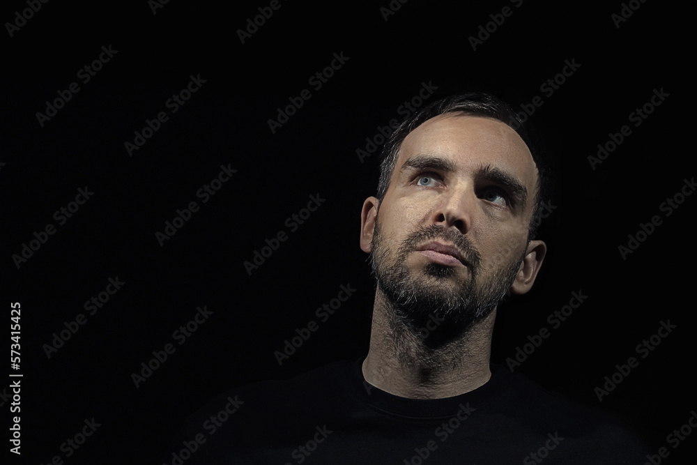 Portrait of a serious man with a small beard, looking thoughtfully to the side. A mid-aged Caucasian man on a black background wearing a black shirt.