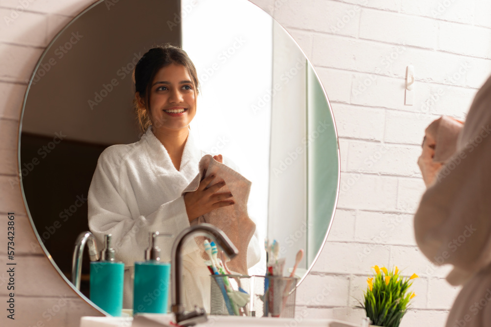 Cheerful woman in bathrobe looking herself in bathroom mirror while wiping hands on towel Cheerful woman in bathrobe looking herself in bathroom mirror while wiping hands on towel