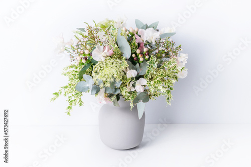 Beautiful flower arrangement of delicate pink flowers, Eucalyptus leaves, Queen Anne's Lace, and lush green foliage, in a grey vase, with a white background.