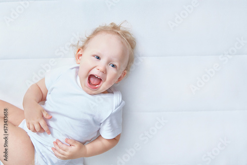 Portrait of cute child. Fun happy laughing baby on a white mattress in white bedroom. Textile and bedding for kids. Kid looking into the camera. Happy family. Close up.