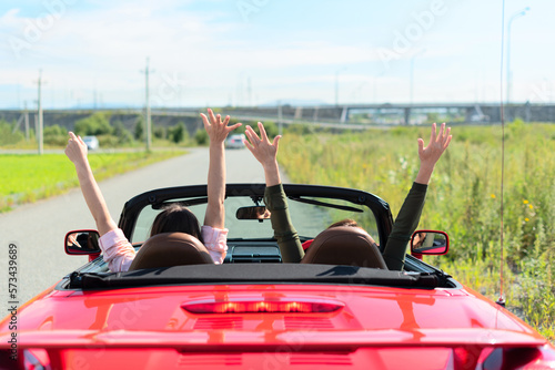 Back view of happy girls driving red cabriolet car with raised hands during vacation road trip having fun together discovering new places. Road trip travel enjoying freedom concept. Selective focus