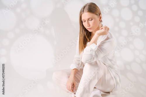 Thoughtful young woman sitting on floor with her head in light light clothes isolated on white background with light art shadows