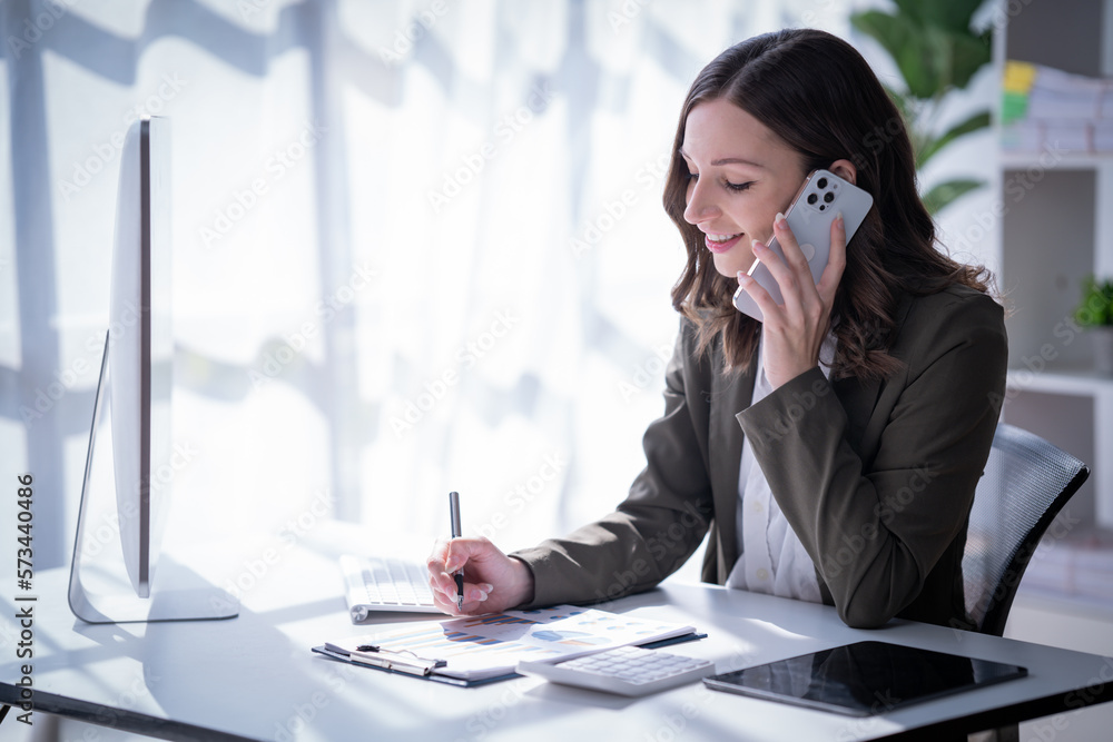 Sharing good business news. Attractive young businesswoman talking on the mobile phone and smiling while sitting at her working place in office and looking at laptop PC. Sharing good business news. Attractive young businesswoman talking on the mobile phone and smiling while sitting at her working place in office and looking at laptop PC.