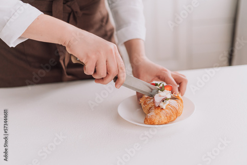 Confectioner in brown apron cuts croissant with knife.