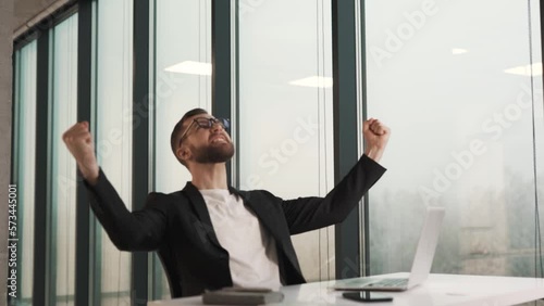 A happy man in business clothes in the office looks at the laptop screen, raising his hands photo