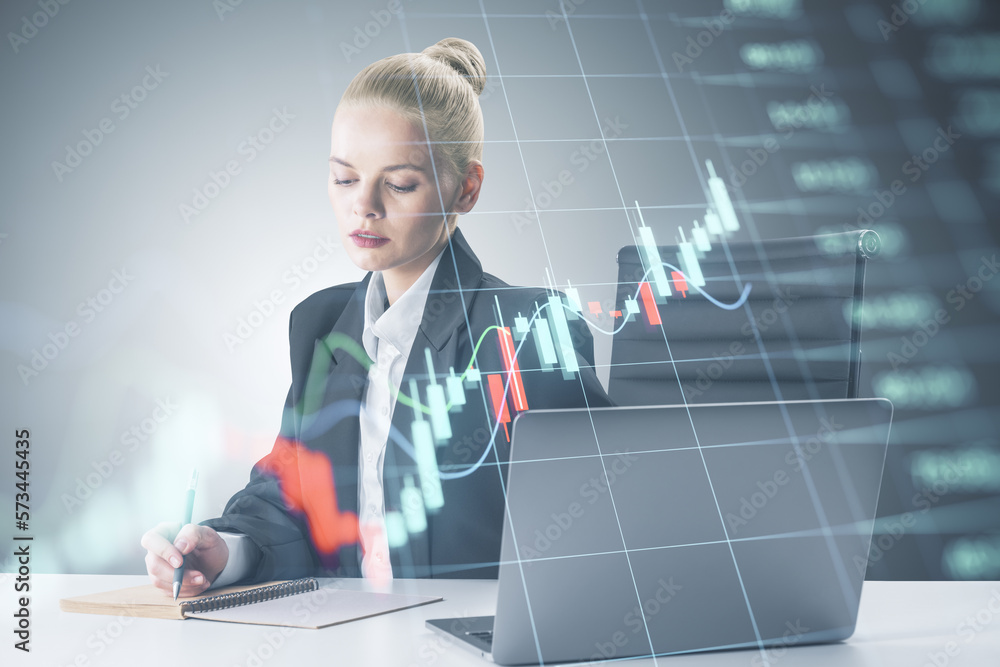 Attractive young european businesswoman at desktop using laptop and writing with forex chart on blurry background. Worker, success, market, trade and invest concept. Attractive young european businesswoman at desktop using laptop and writing with forex chart on blurry background. Worker, success, market, trade and invest concept.