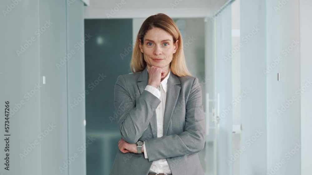 Middle aged confident focused business woman in formal suit standing in modern light office puts hand on chin smile looks at camera feels happy. 40s pretty positive female face portrait concept. Middle aged confident focused business woman in formal suit standing in modern light office puts hand on chin smile looks at camera feels happy. 40s pretty positive female face portrait concept.