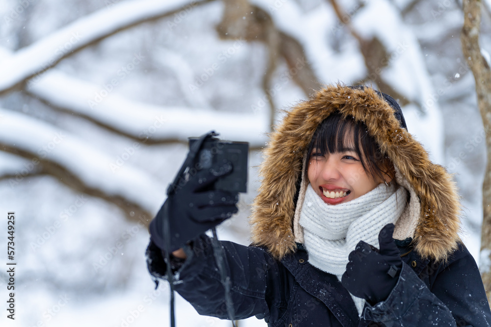Asian woman using digital camera taking selfie during walking down small town street covered in snow. Attractive girl enjoy travel local village near the mountain in Japan on winter holiday vacation.