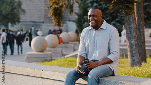 Happy smiling African American ethnic middle-aged man businessman male sitting outdoors in city with coffee tea and phone waiting for date business meeting smile to friend carefree cheerful relaxing