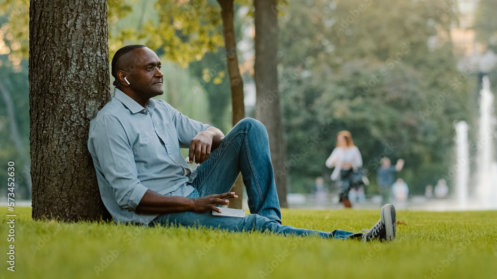 Obraz premium Calm relaxed sad serious middle-aged male listening music with earphones mature adult African American ethnic man in wireless headphones sitting on grass in city park enjoy audio song sound outdoors