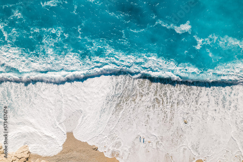 Bird's-eye view of a couple of people on a sandy beach with turquoise clear water with foaming big waves. Drone shot top view.