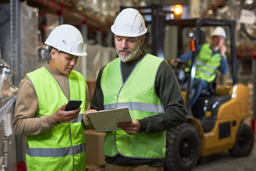 Waist up portrait of two workers holding clipboard in warehouse and wearing reflective jackets Waist up portrait of two workers holding clipboard in warehouse and wearing reflective jackets