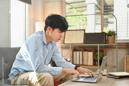 Asian male freelancer communicating with client via laptop computer while sitting on couch in home office