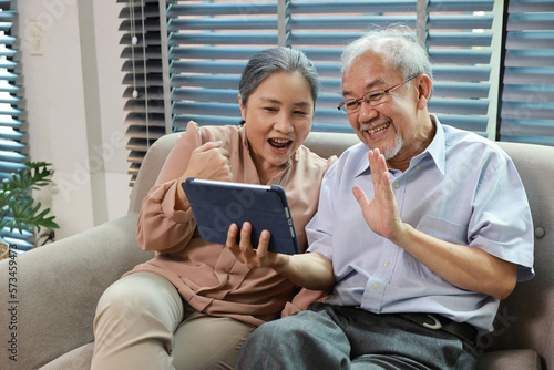 Happy smiling asian senior couple sitting on sofa and using tablet while online video call with friend or relative cousin at home living room. Internet information technology and lifestyle concept.
