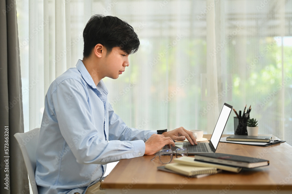 Side view of asian man freelancer using laptop computer and working marketing project at bright home office Side view of asian man freelancer using laptop computer and working marketing project at bright home office
