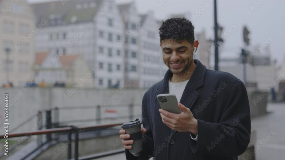 Multi-Ethnic Man Wearing a Coat, Smiling, and Walking Outside While Using Earphones and Smartphone. Winter Time. People and Technology Concept Multi-Ethnic Man Wearing a Coat, Smiling, and Walking Outside While Using Earphones and Smartphone. Winter Time. People and Technology Concept