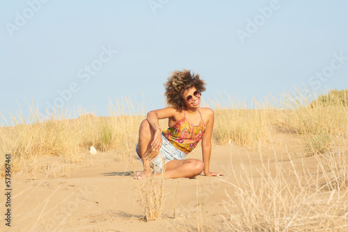 Pretty girl with afro in sunglasses sitting on sand. Portrait of happy beautiful woman with dark brown frizzy hair wearing sunglasses and top with shorts, sitting on sand in desert on sunny day.