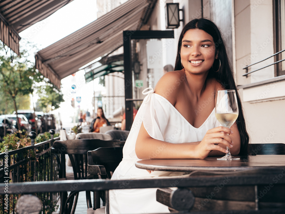 Young beautiful smiling hipster female in white dress. Sexy carefree woman sitting at cafe in the street. Positive model drinking white wine. Cheerful and happy. Outdoors at sunny day