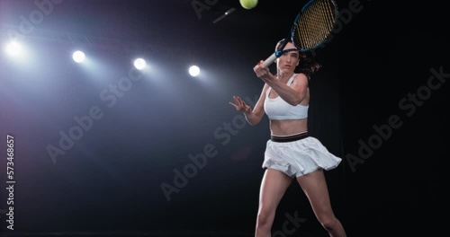 Aesthetic Shot of an Athletic Female Tennis Player with a Black Background Hitting a High Ball Under Spotlights. Arc, Cinematic Super Slow Motion Captures a Winning Strong Shot During a Championship photo