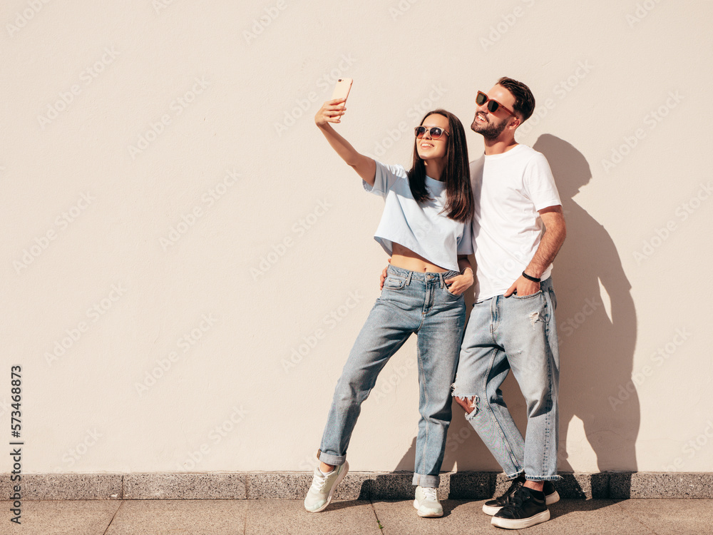 Smiling beautiful woman and her handsome boyfriend. Woman in casual summer jeans clothes. Happy cheerful family. Female having fun. Sexy couple posing in street near wall. In sunglasses. Takes selfie