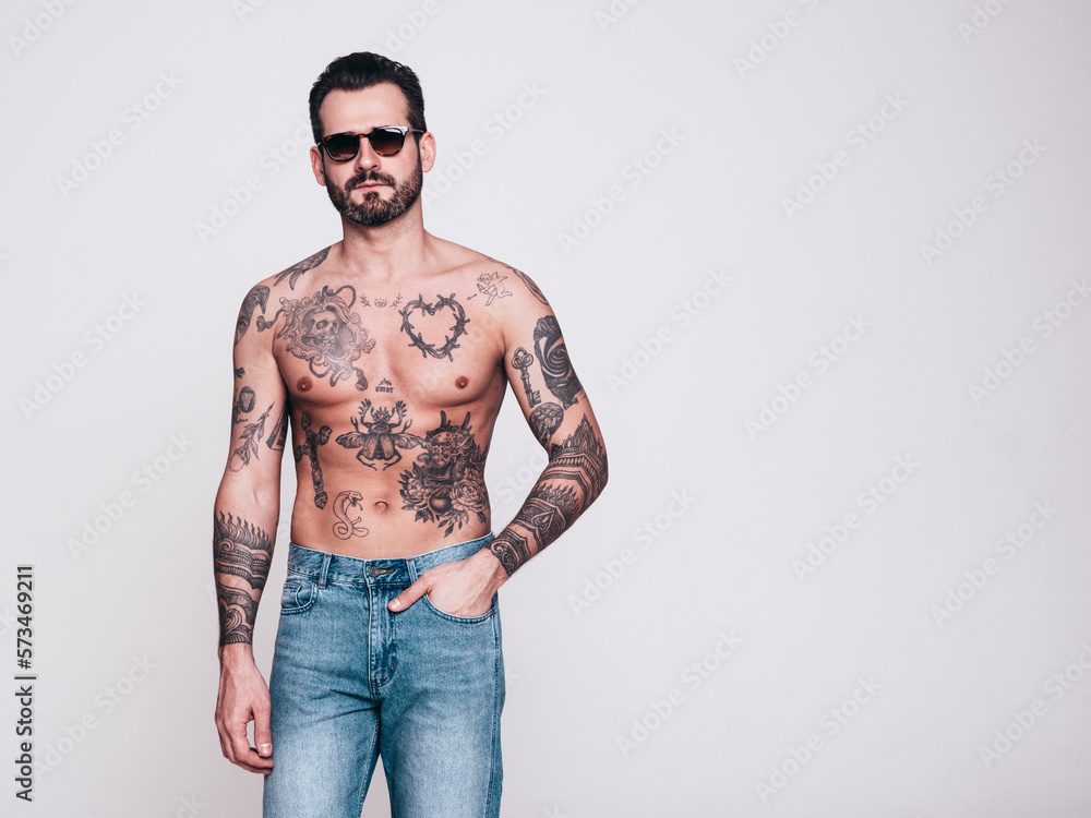 Fototapeta premium Portrait of handsome confident stylish hipster lambersexual model in jeans. Sexy modern man. Naked torso with tattoos. Fashion male posing in studio. Isolated on white. In sunglasses