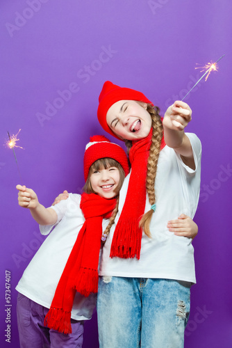 Pair of Happy Winsome Embracing Exclaiming Caucasian Girlfriends in Seasonal Warm Clothing Having Fun With Bright Burning Bengal Lights On Lifted Hands On Purple.