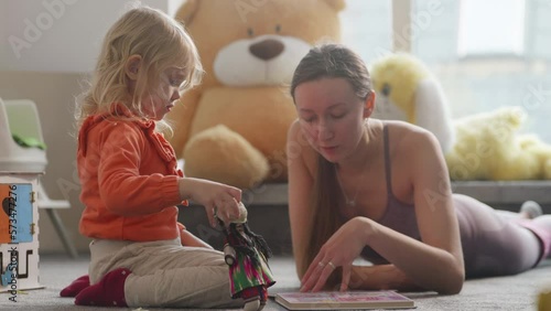 Young sporty mom teaches her daughter with a book on the floor. Cute blond kid, pretty women wearing bright clothes, nursury room filled with natural light. Finding time for sports being a parent. 4K