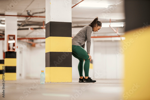 An exhausted sportswoman in shape is leaning on her knees and resting from workouts in garage.