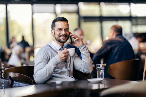 A young happy man in smart casual is sitting in cafe and drinking coffee while having a phone call.