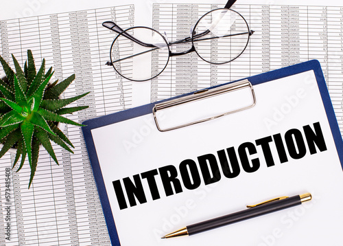 Folded business papers, pen, glasses, a flower in a pot and a tablet with a sheet of paper with the text INTRODUCTION on the desktop. Top view of the workplace. Business concept