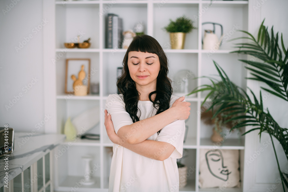 Portrait of a young brunette woman hugging herself in a bright stylish room. Beautiful brunette girl wrapped her arms around herself Portrait of a young brunette woman hugging herself in a bright stylish room. Beautiful brunette girl wrapped her arms around herself