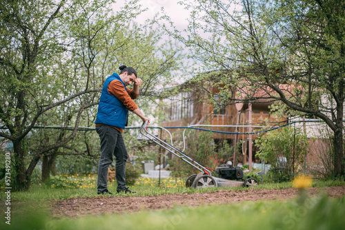 Portrait of a young farmer, gardener with a lawn mower on a plot of land in spring.