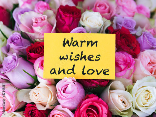 A yellow greeting card with words Warm Wishes and Love laying on a bouquet of colorful roses