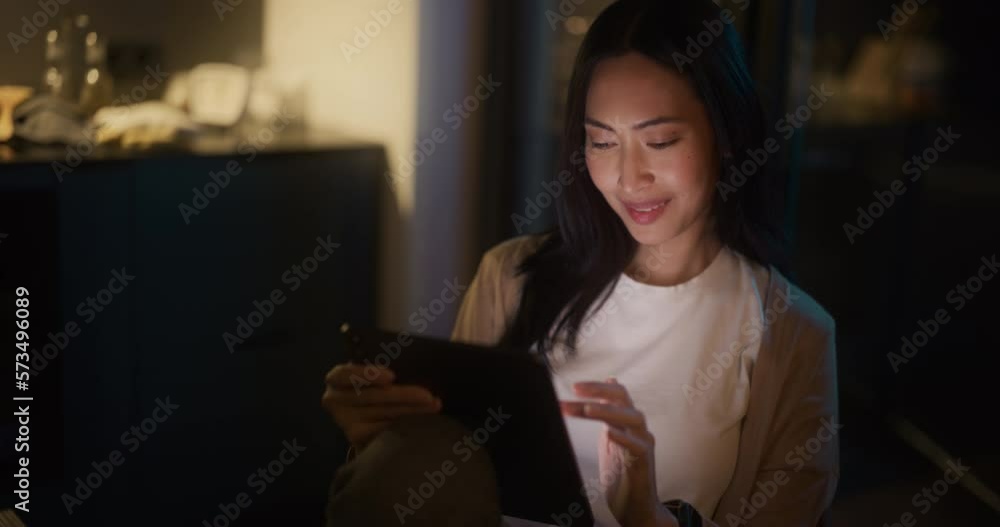 Beautiful Authentic Asian Woman Sitting at a Table in Cozy Kitchen and Using Digital Tablet at Home at Night. Female Smiling and Checking Social Media, Doing Online Shopping, Watching Internet Videos Beautiful Authentic Asian Woman Sitting at a Table in Cozy Kitchen and Using Digital Tablet at Home at Night. Female Smiling and Checking Social Media, Doing Online Shopping, Watching Internet Videos