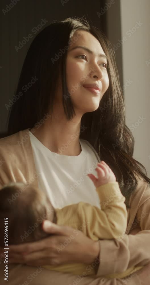 Vertical Screen: Portrait of an Asian Mother Holding her Baby in her Arms and Looking at the Street Through Window at Home. Calm Woman Enjoying the Sunlight, Feeling Warm and Safe with her Toddler Vertical Screen: Portrait of an Asian Mother Holding her Baby in her Arms and Looking at the Street Through Window at Home. Calm Woman Enjoying the Sunlight, Feeling Warm and Safe with her Toddler