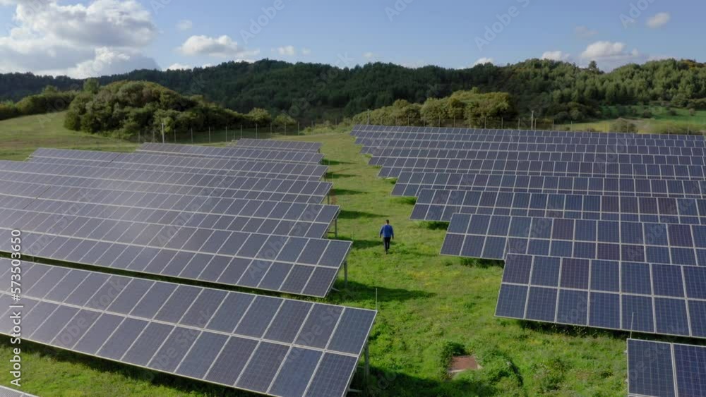 Aerial: slow pan of one man walking between many rows of solar panels in a green field during a sunny day Aerial: slow pan of one man walking between many rows of solar panels in a green field during a sunny day