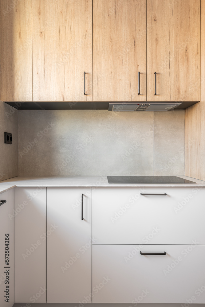 Part of kitchen with a table along wall. Table with gray marble top and black hob. Above stove there is ventilation system built into cabinet. Daylight pours in from window, illuminating kitchen. Part of kitchen with a table along wall. Table with gray marble top and black hob. Above stove there is ventilation system built into cabinet. Daylight pours in from window, illuminating kitchen.
