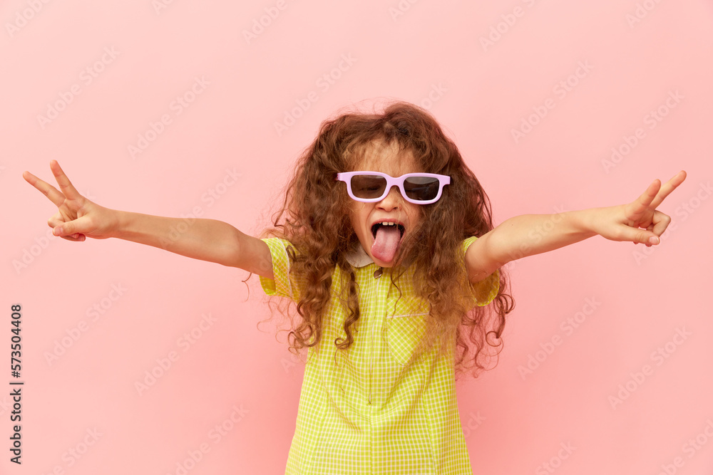 Tongue out. Little emotional girl, child with curly hair posing in yellow dress and sunglasses over pink studio background. Concept of childhood, emotions, fun, fashion, lifestyle, facial expression Tongue out. Little emotional girl, child with curly hair posing in yellow dress and sunglasses over pink studio background. Concept of childhood, emotions, fun, fashion, lifestyle, facial expression