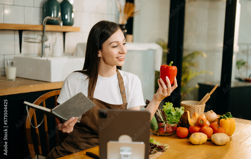 Young beautiful woman in the kitchen in an apron, fresh vegetables on the table, writes down her favorite recipes, comes up with ideas for dishes.