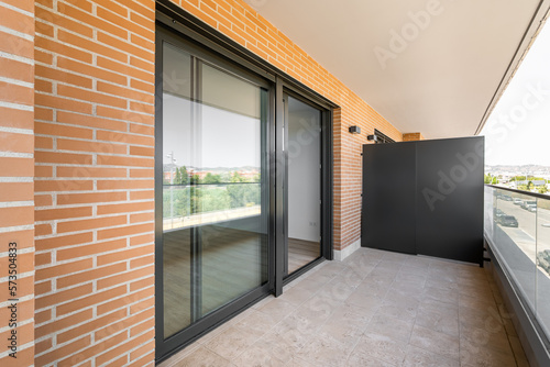 Large entrance balcony door made of black plastic with a sliding system. Terrace with marble floor and glass railings. The walls of the building are made of yellow bricks.