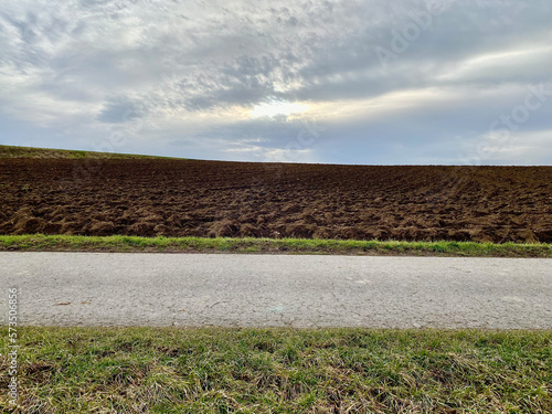 Landstraße seitlich vor Landschaft mit Grünstreifen, Straße, Grünstreifen, braunen Acker und Himmel mit Wolken