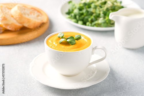 Cup of pumpkin cream soup garnished with fresh sunflower microgreen. Creamy butternut squash autumn soup in elegant white tea cup.  Front view, selective focus