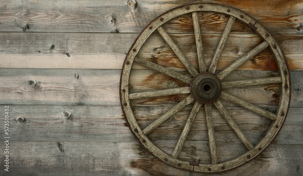 An old wooden carriage wheel with metal fittings hangs on the wall of a wooden house for decoration. Wooden spoke wheel. Outdoor natural light. An old wooden carriage wheel with metal fittings hangs on the wall of a wooden house for decoration. Wooden spoke wheel. Outdoor natural light.