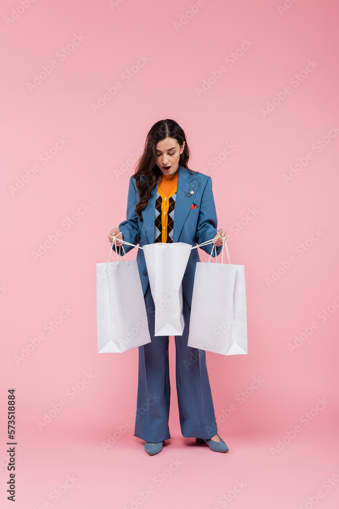 full length of surprised young woman in blue pantsuit looking into shopping bag on pink.