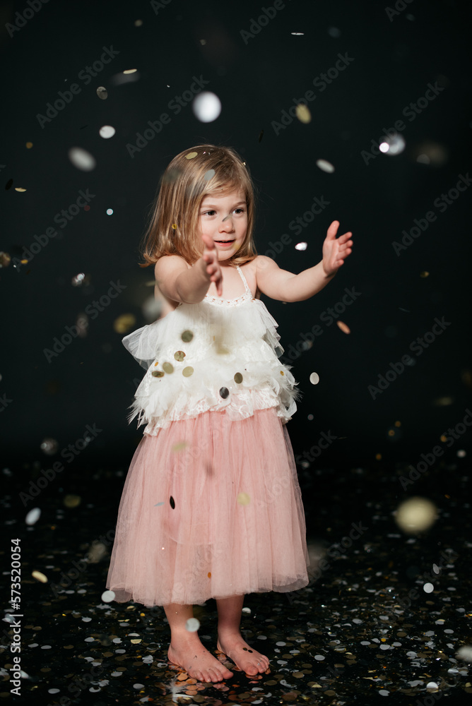 Fototapeta premium little girl in a dress playing with confetti against black backdrop