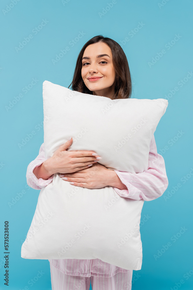 pleased young woman in sleepwear holding white pillow isolated on blue. pleased young woman in sleepwear holding white pillow isolated on blue.