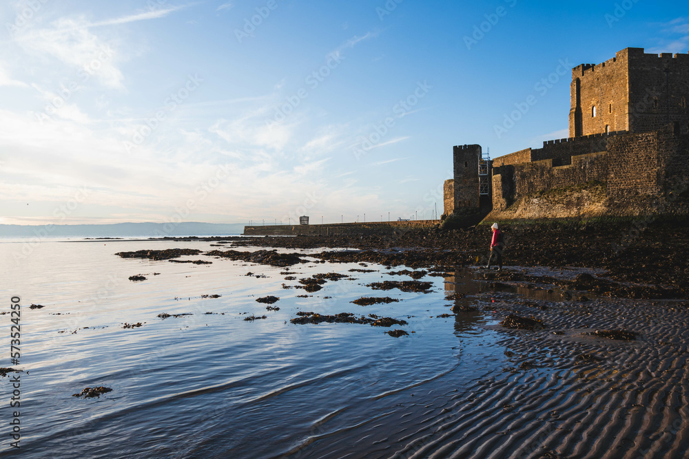 Silhouette of Carrickfergus Castle, Northern Ireland, wide angle lens with sunrise, blue sky, water reflections and horizon with a woman walker in winter red jacket and hat